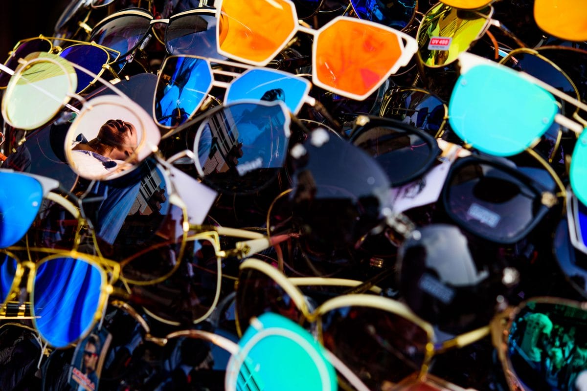 A pile of sunglasses with a woman reflected in some of the lenses.