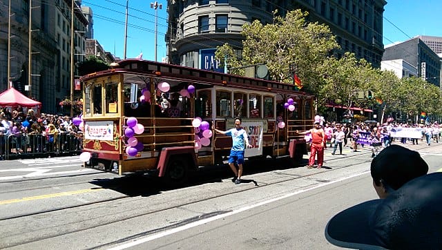 Tram with balloons on it with parade of people walking by in street.