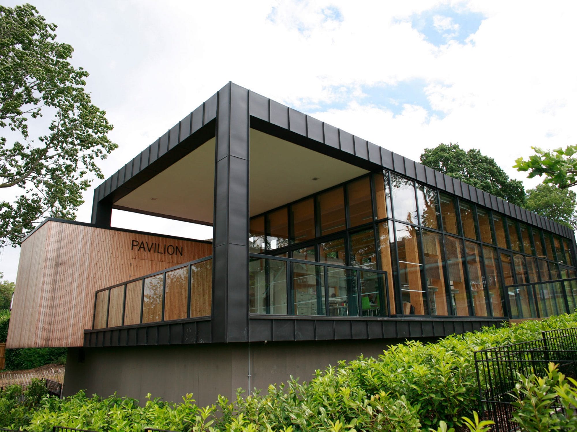 A square building as seen from outside and slightly below at an angle. The end closest to the camera is open to a balcony, with a glass walled internal room behind it. There are trees and shrubs surrounding it