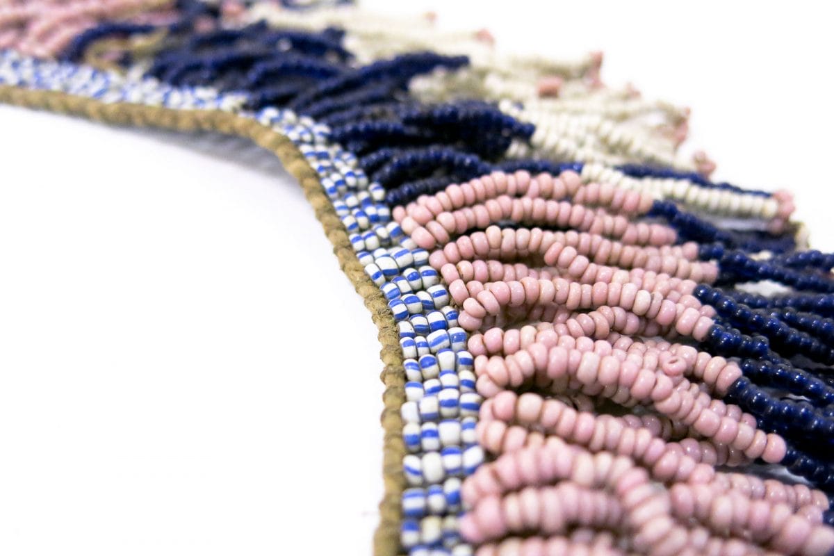 A rope of beads, white, pink and blue, strung together in a neck piece. They are seen in close up against a white backdrop