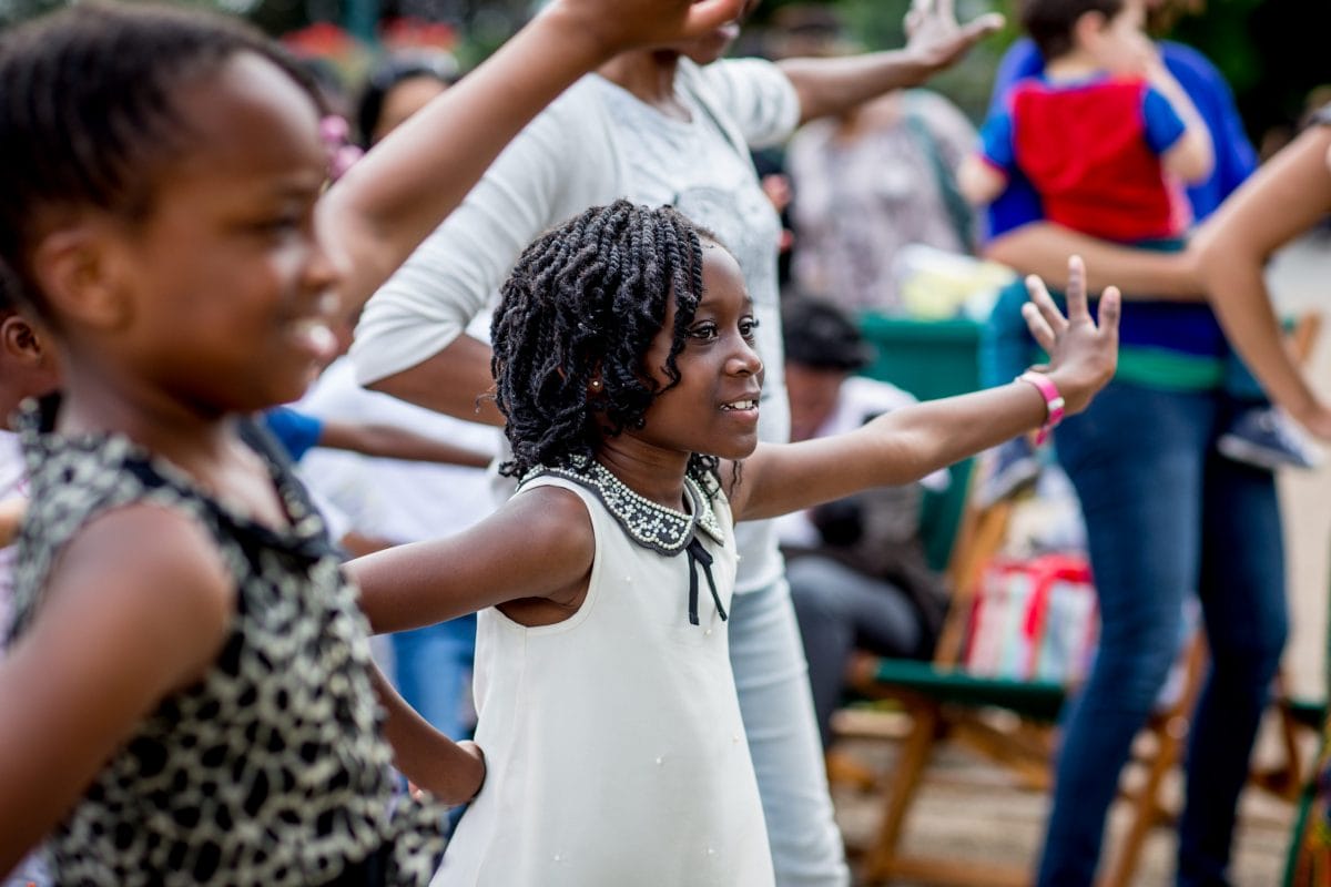 Two girls in close up, in the middle of doing a dance pose, in among other adults and children dancing - they are smiling and their arms are in the air