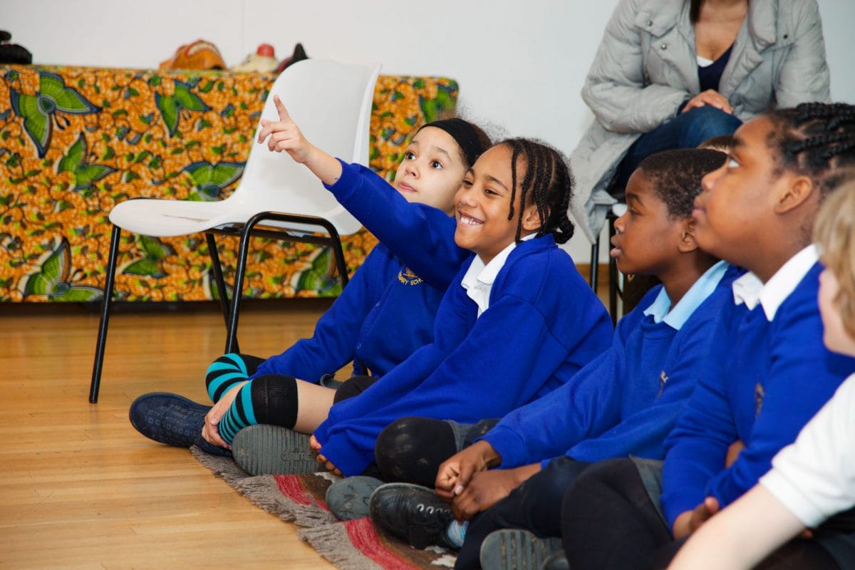 A row of children in blue jumpers sit cross legged on a wooden floor, smiling and raising their hands