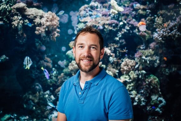 A man in a blue top is seen from the chest up in front of a large aquarium stank with small fishes and corals in it