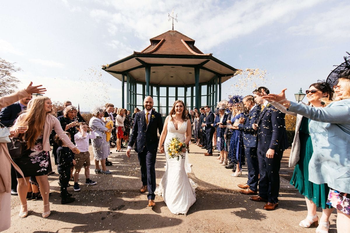 A bride and groom walk in front of the Horniman bandstand as guests throw confetti