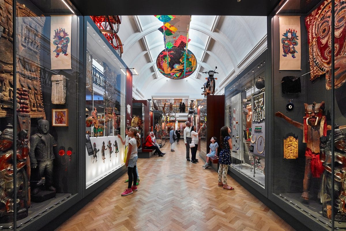 A corridor in a gallery with people looking into cases. There are kites on the ceiling