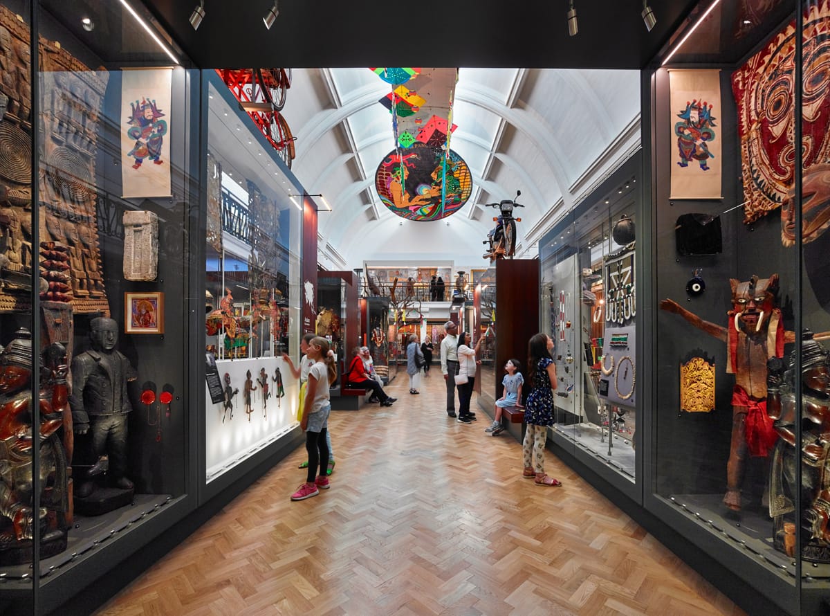 A corridor in a gallery with people looking into cases. There are kites on the ceiling