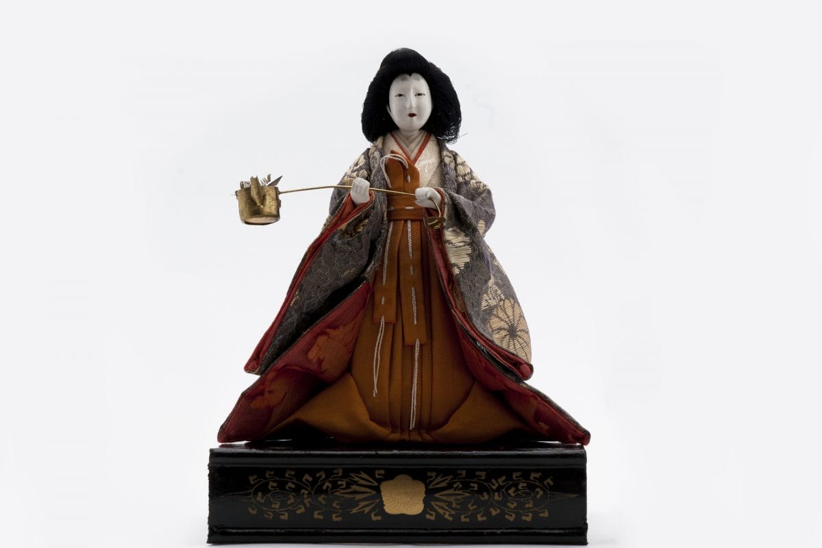 Figure of doll with painted white face and long wide dress. The doll is holding a small pot on a stick and the doll is stood on a black box with gold detailing.