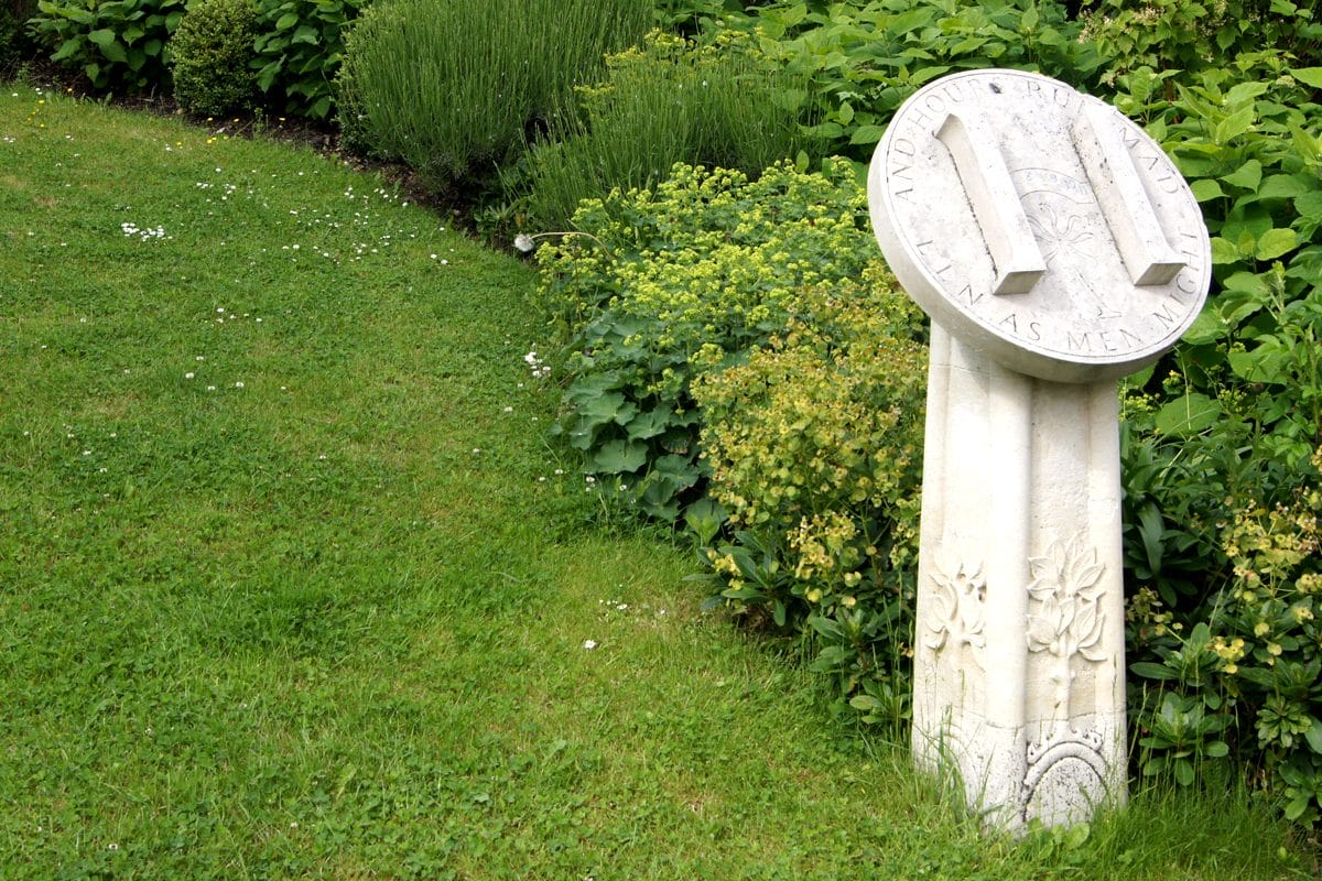 A stone sundial in a garden with a H on the front