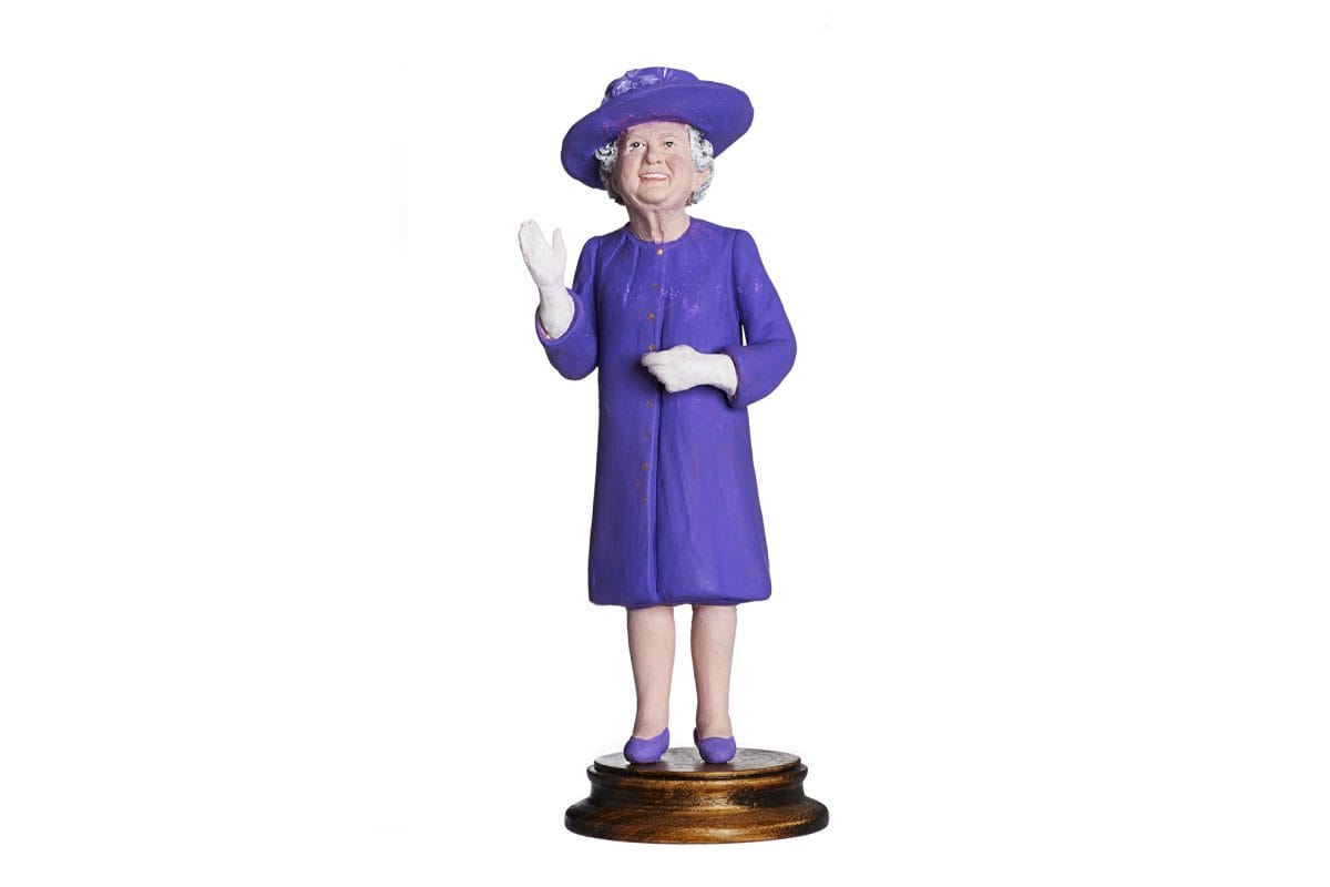 Figure of the Queen wearing purple hat, shoes and skirt suit. Figure mounted on a wooden stand.