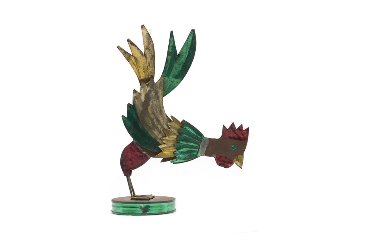 Figure in the shape of a cockerel made from sheets of metal and partially decorated with metallic paints in red, yellow and green. The figure is standing on a hollow, circular metal pedestal.