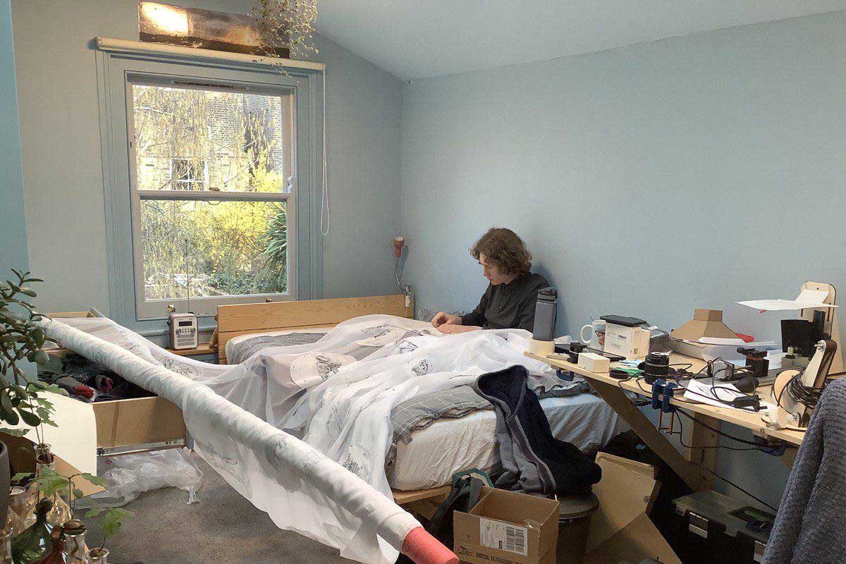 A person sits on a bed stitching a huge net curtain