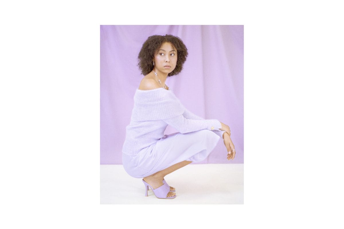 A Black woman with short curly hair wearing a purple midi dress and purple heeled mules sits on her haunches, and looks slightly to her right