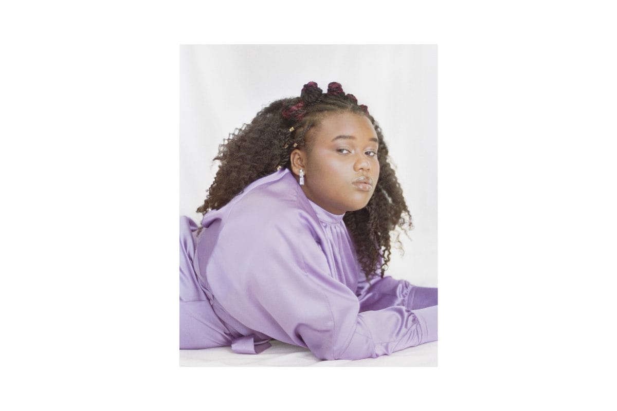 A Black woman lies on her front and looks at the camera which is on her right. She is wearing a purple high necked dress, and has mid length curly hair, some of which is braided