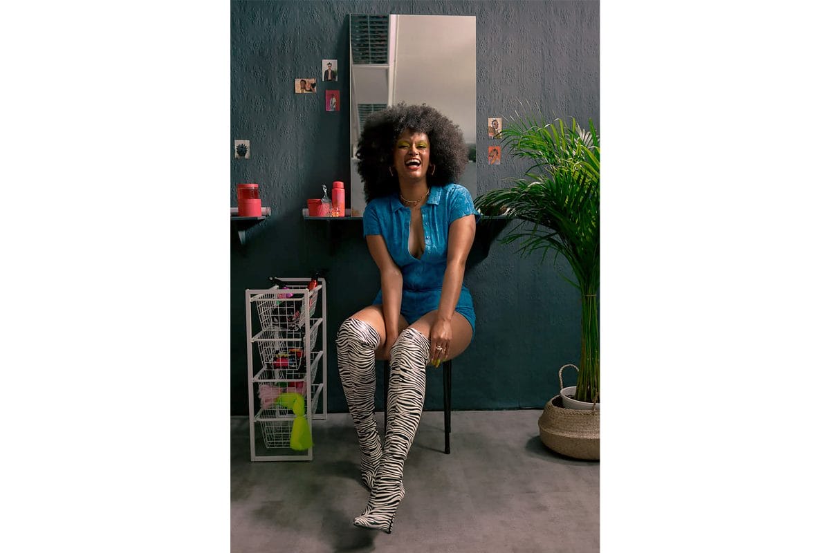 A Black woman with afro hair sits on a chair in a salon and laughs as she looks at the camera. She is wearing a blue playsuit and thigh high zebra print boots which she is pulling up