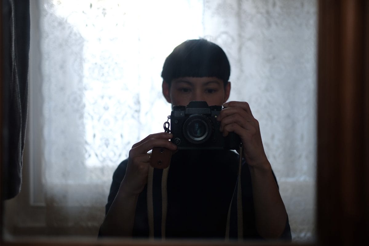 A woman takes a photo in a mirror with a camera. We can see the top half of her face, including her bowl cut hair