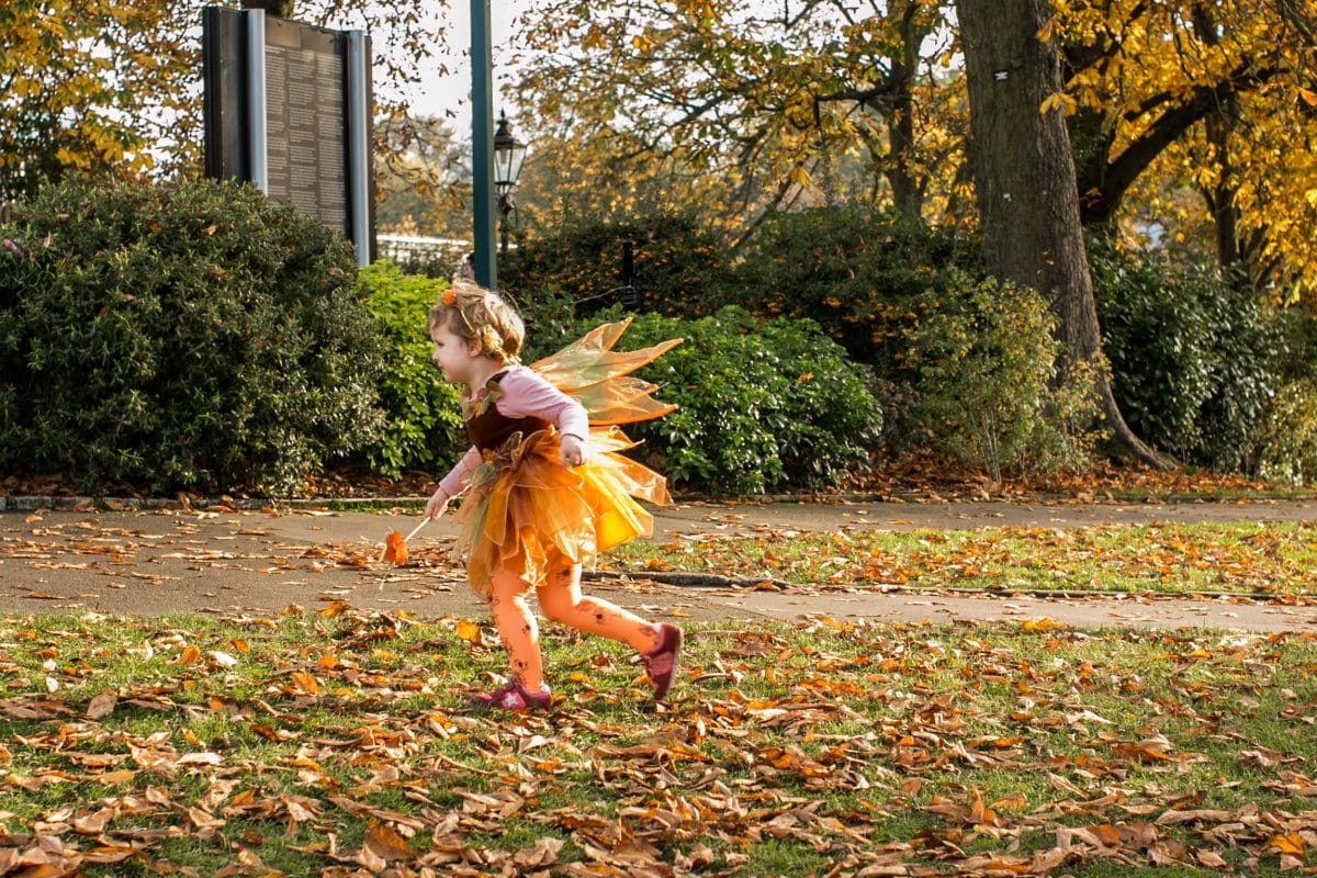A girl in an orange fairy dress runs through leafy grass at the Horniman