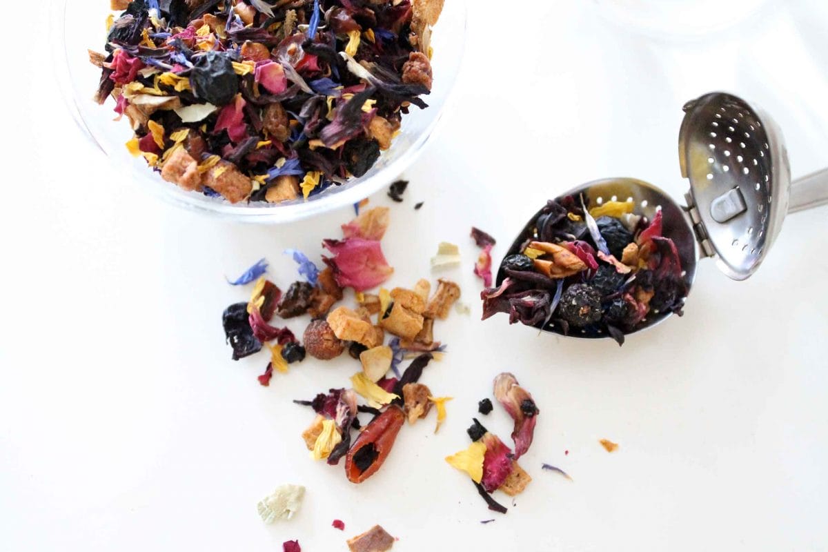 A bowl of tea leaves and a tea strainer filled with tea leaves sit next to one another on a surface strewn with tea leaves