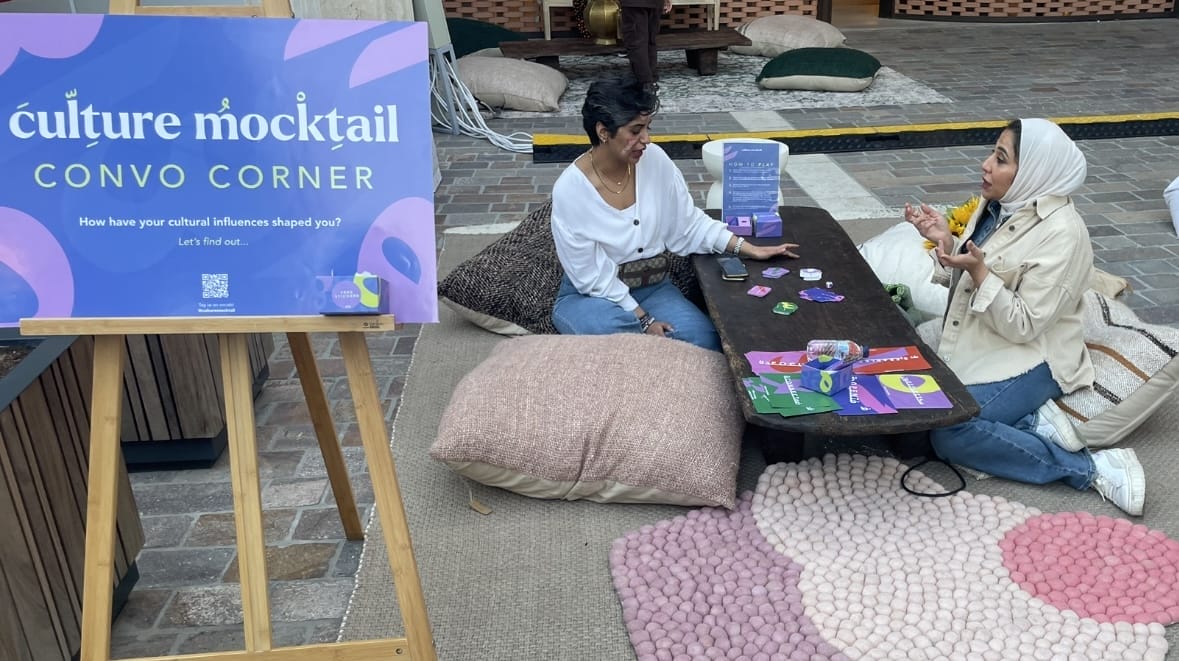 Two people sat on the floor and talking across a low table. Sign next to them reads 'culture mocktail convo corner'