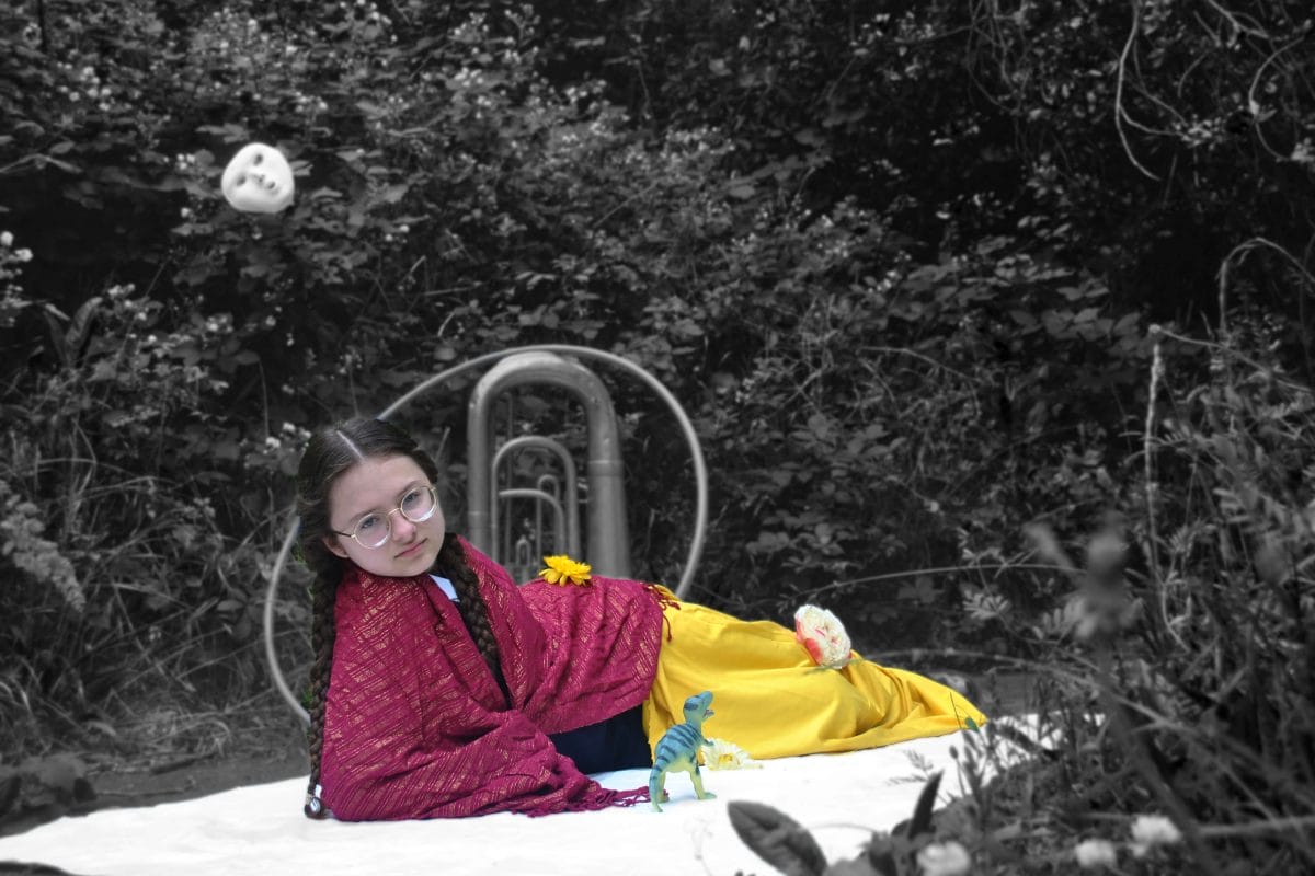 A girl lies on a white blanket outside, wearing a red shawl and a yellow skirt. She is in colour and her surrounding are in black and white. She is looking at the camera.