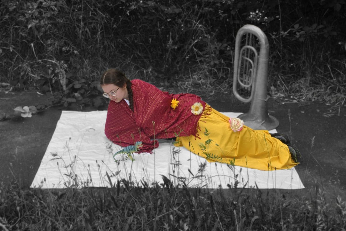 A girl lies on a white blanket outside, wearing a red shawl and a yellow skirt. She is in colour and her surroundings are in black and white. She is looking at the camera. She looks down at the white blanket.