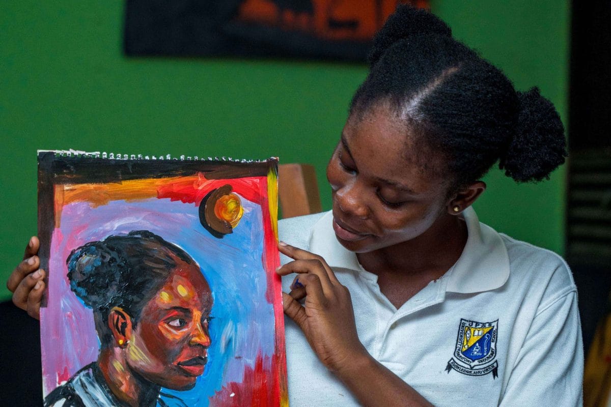 A girl holds up and looks at a self portrait she has painted