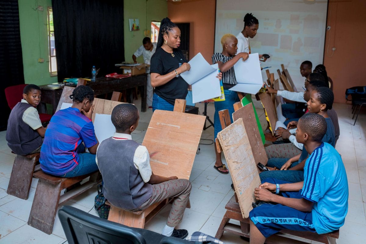 Students with easels are gathered in a circle around a teacher holding a folder of paper