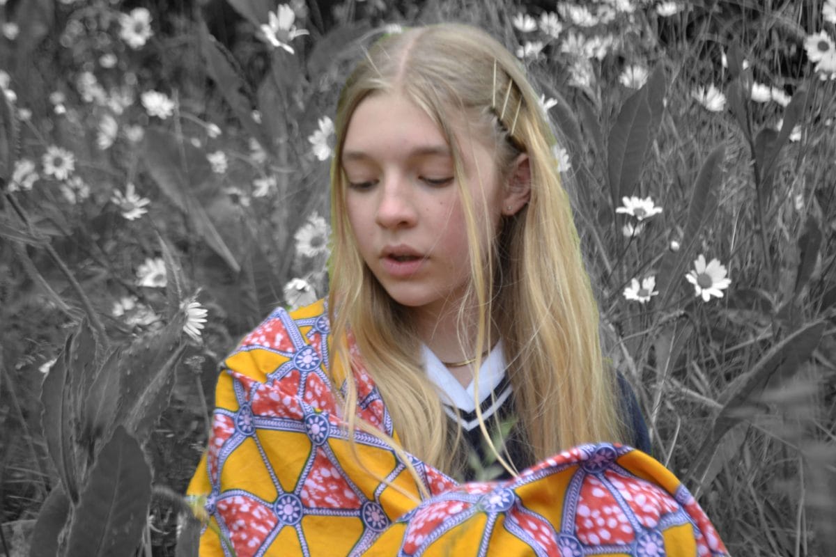 A girl with blonde hair is wearing a colourful patterned shawl. She is in colour and her surroundings of long grass and flowers are in black and white.