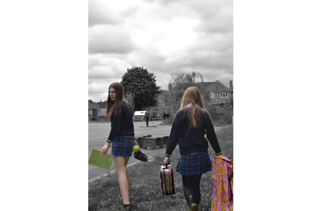 Two girls walk across some grass to a playground. They are carrying a suitcase, some material, some paper and a tennis ball. One of the girls is half turned to look at the camera. They are in colour and their surrounding are in black and white.