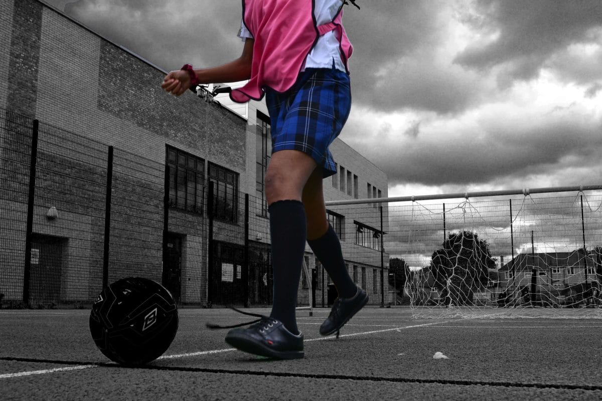 The bottom half of a child plays football, wearing a pink bib and with school playground behind. They are in colour and their surroundings are in black and white.