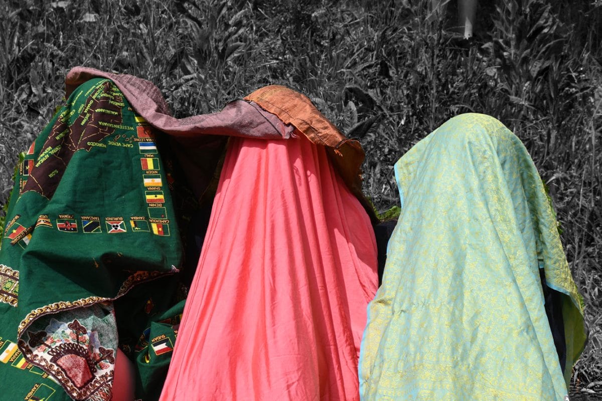 Three girls stand in a row with colourful material over their heads, covering them completely. They are in colour and their surroundings are in black and white,