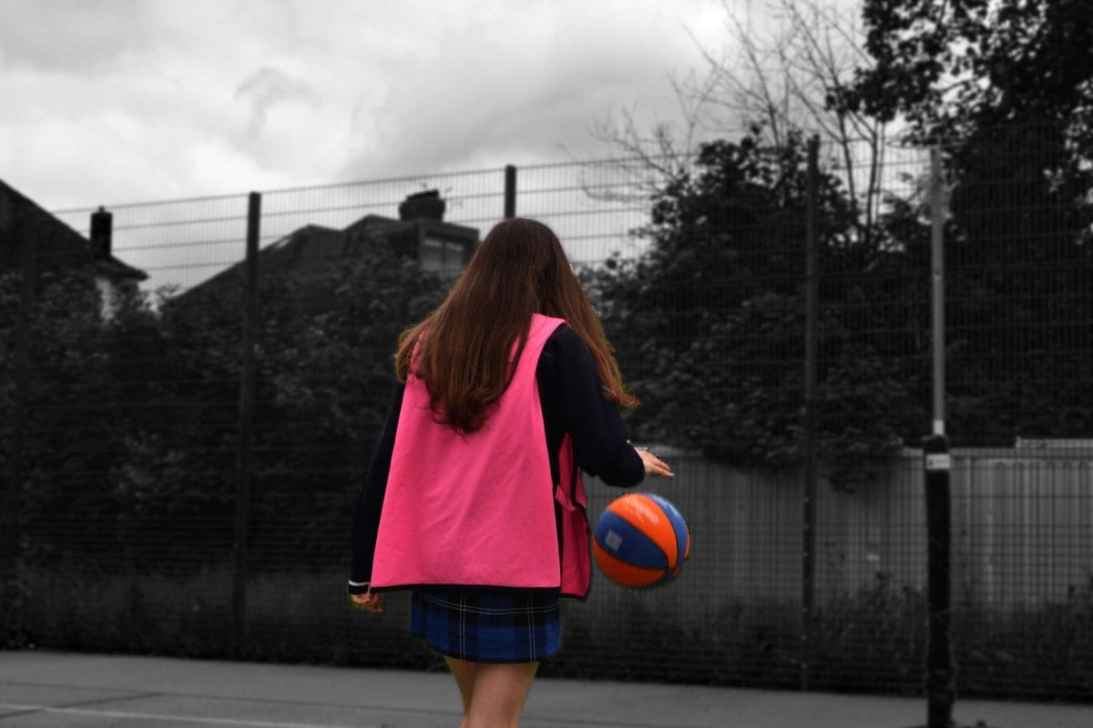 The back of a girl bouncing a basketball on a court, wearing a pink bib. She is in colour and her surroundings are in black and white.