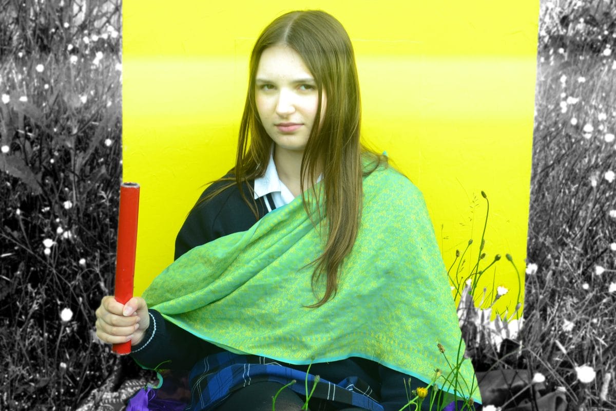 A girls sits amongst grass against a yellow background and wearing green material across her shoulder. She holds up a red pipe. She is in colour and her surroundings are in black and white