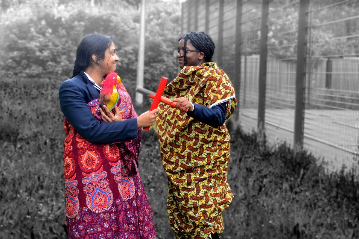 Two girls wear colourful patterned material wrapped around them and are each holding a red pipe which are touching. One of the girls is holding a parrot model. They are in colour and their surroundings are in black and white.