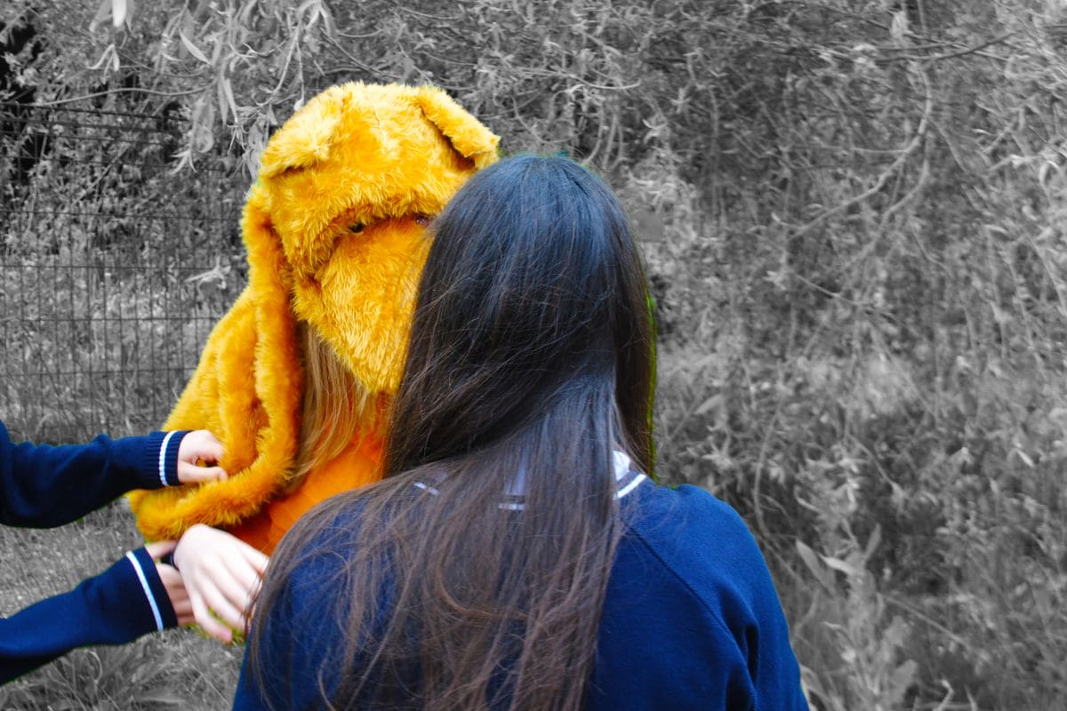 The back of a girls head faces a large yellow teddy bear's head, with trees and bushes surrounding. The girl and the bear's head are in colour and the surroundings are in black and white