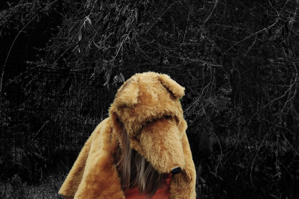 A child with a teddy bear's head stands amongst trees. They are in colour and their surroundings are in black and white.