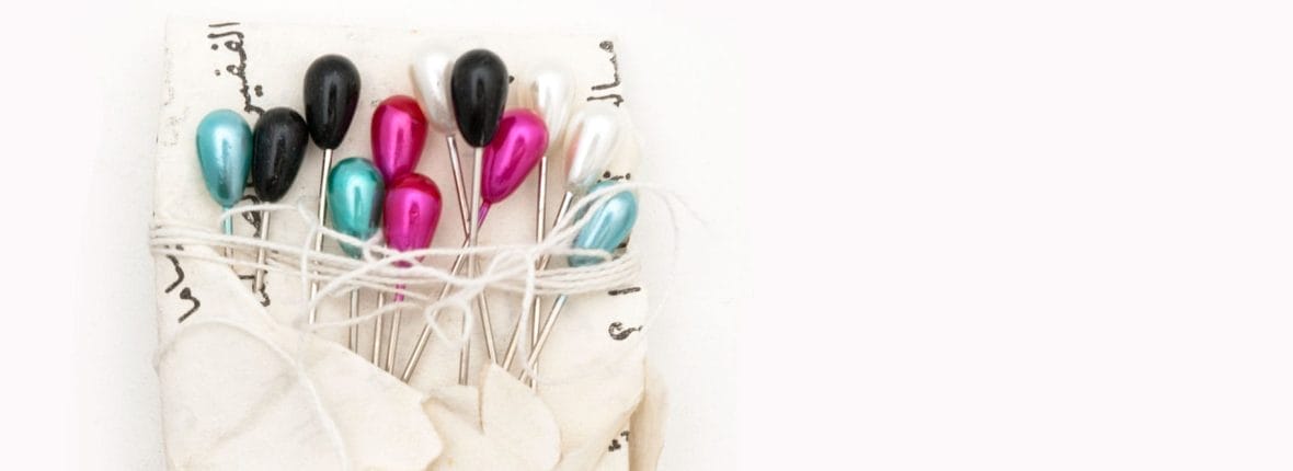 Pins with multicoloured pearly heads wrapped in a piece of paper. The paper has printed text.