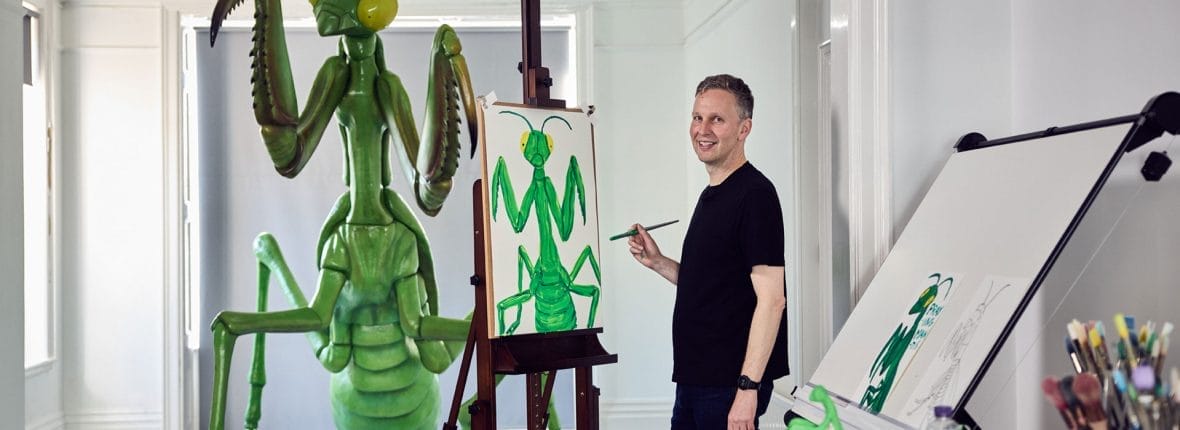 David Shrigley paints a picture of a praying mantis in a studio, behind him is a sculpture of a praying mantis