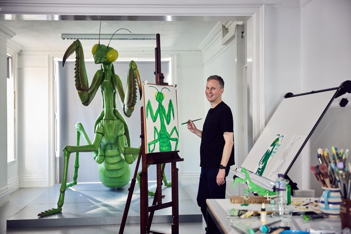 David Shrigley paints a picture of a praying mantis in a studio, behind him is a sculpture of a praying mantis