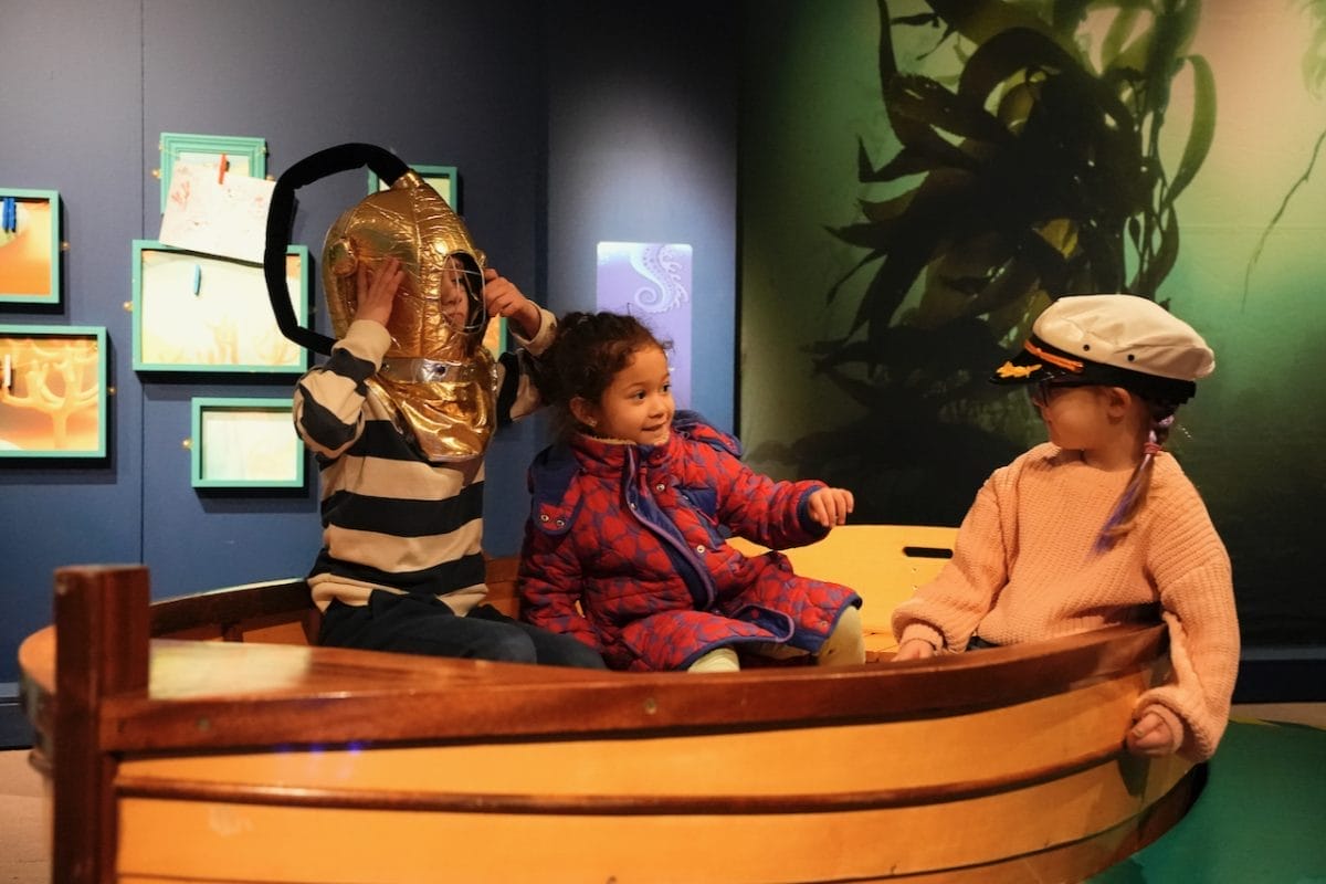 Three children smiling and playing in a wooden boat at the Voyage to the Deep exhibition.