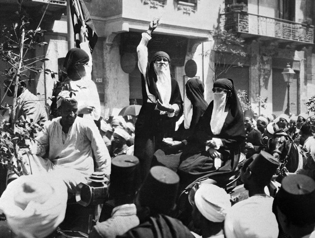 Black and white image of a group of women wearing face coverings and standing amongst a crowd making a speech