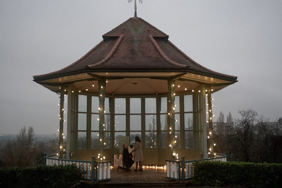 A man proposes to a woman inside a fairy light covered Horniman Bandstand