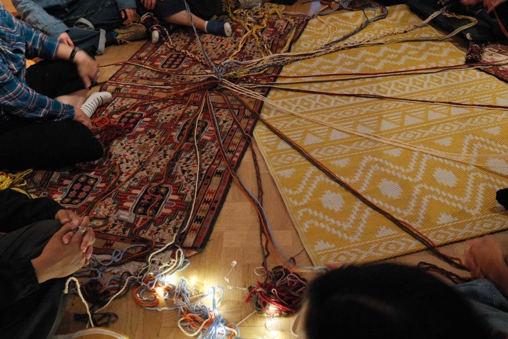 Coloured string in a knot lies on colourful rugs as people sit on floor around it