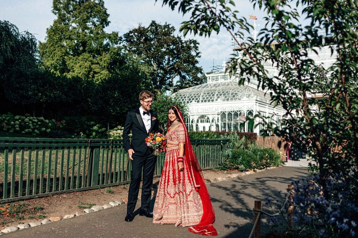 A bride and groom stand amongst greenery with the Horniman Conservatory in the background