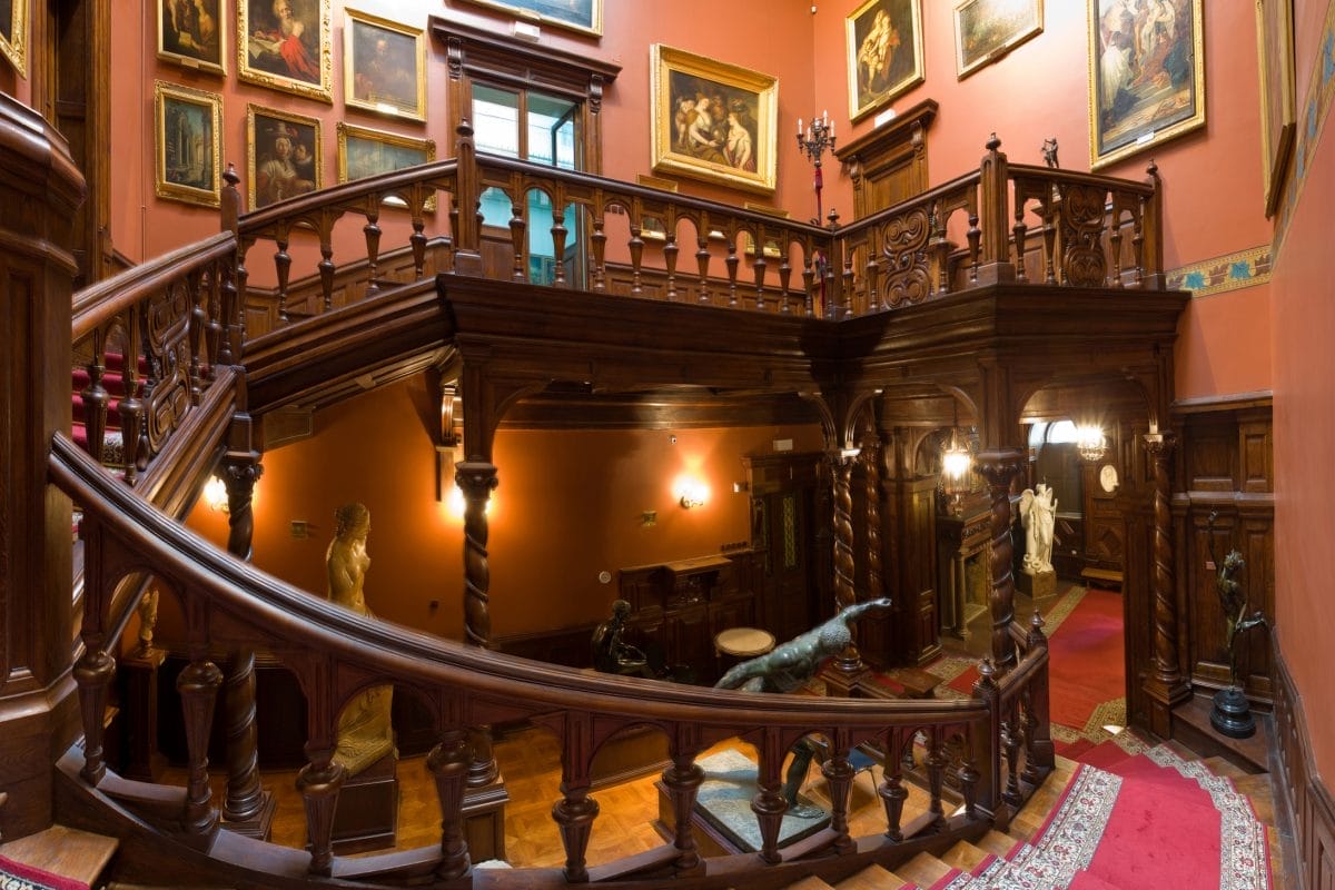 A fish eye lens view of a grand staircase, made of dark wood and carpeted in red. The walls around the staircase show old artworks and statues are dotted around the ground floor
