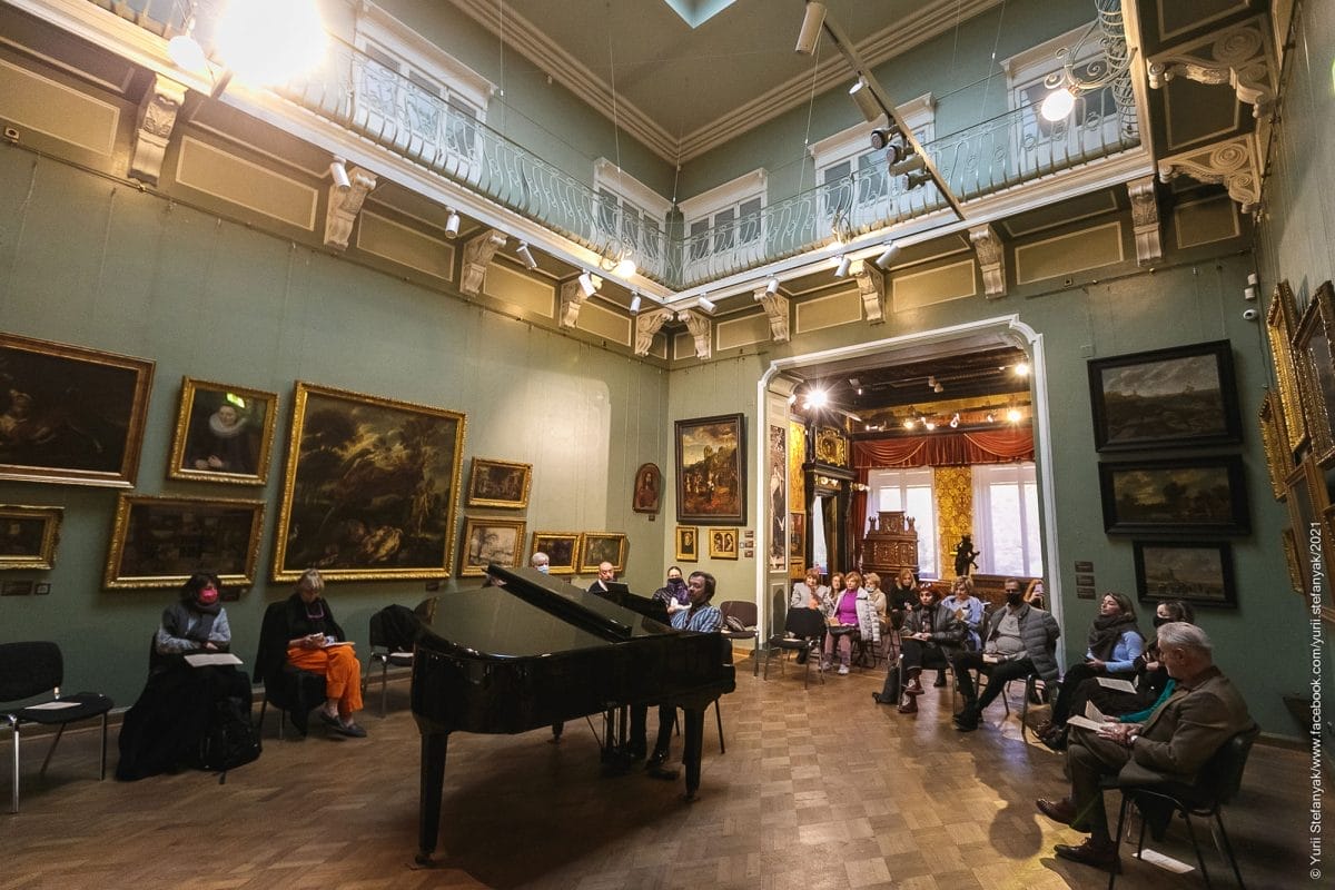 A man plays at a grand piano at the centre of an orante room, with a balcony above the main floor. People gather around to listen, seated. There is old artworks on the walls