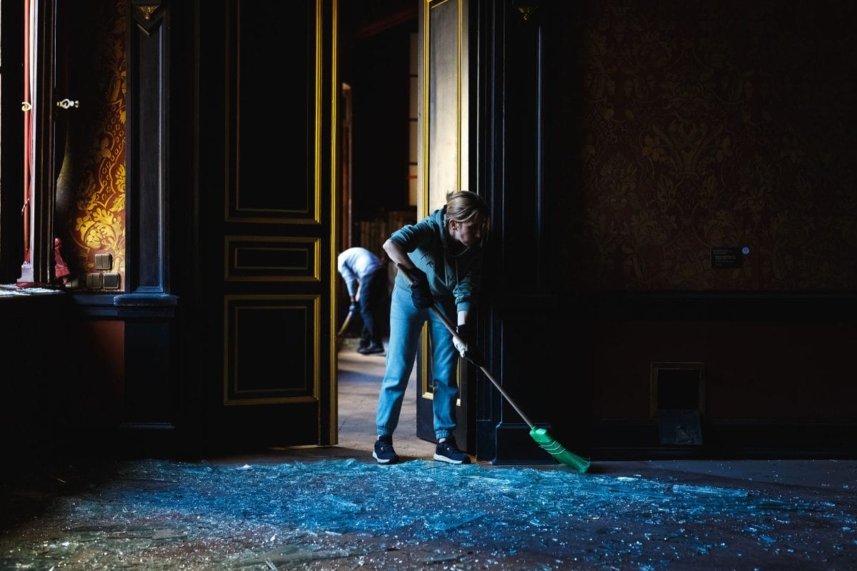 A woman sweeps up glass shattered across the floor with a broom in a dark ornate room