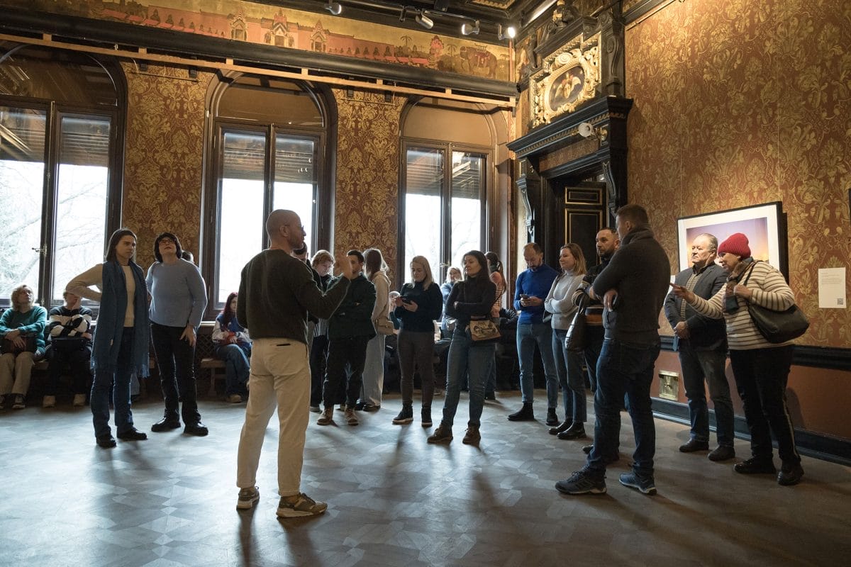 A young man speaks to a group of people in an ornate brown room, with high windows.