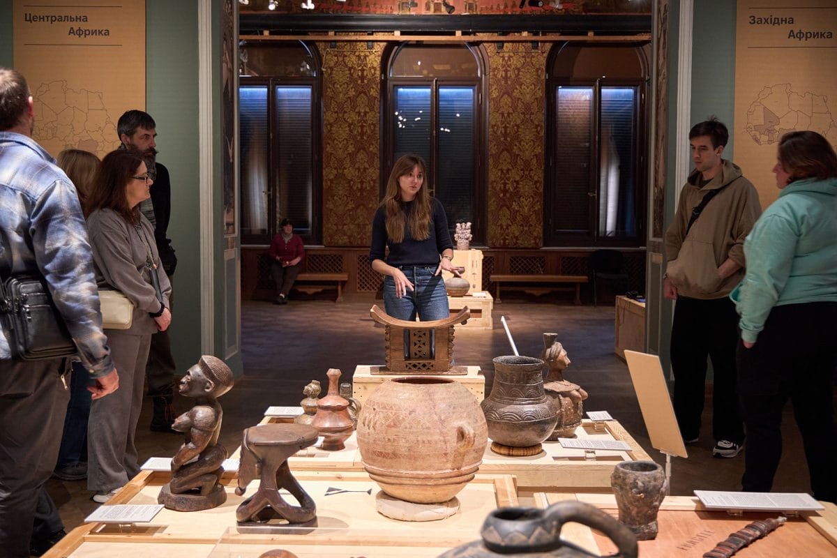 A young woman speaks to a group of people gathered around a collection of objects, which are African in origin - bowls, statues, pots, stools are visible
