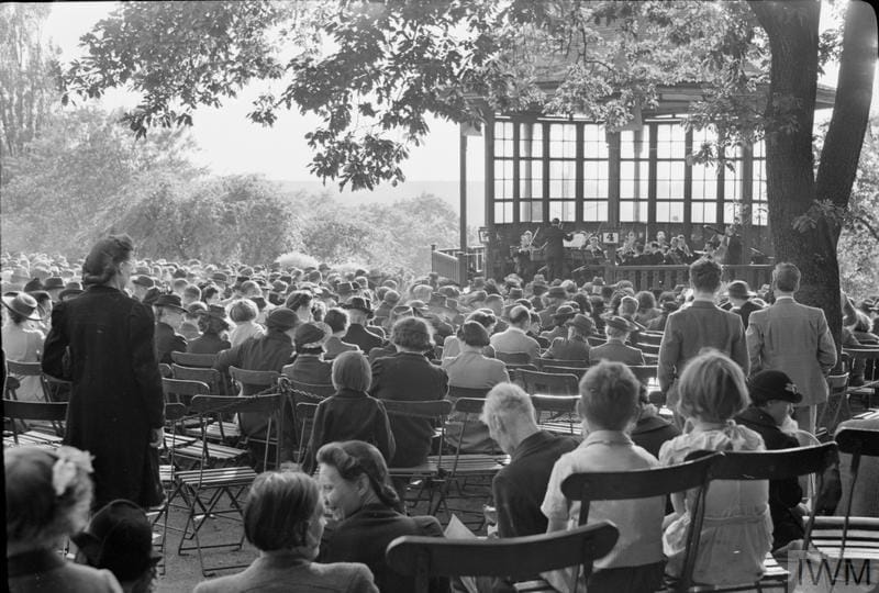 Men, women and children enjoy the last of the Sunday evening sunshine, under the dappled shade of a large tree, as they listen to Jack Gold and his orchestra play 'The Savoy Russian Medley' on the bandstand in the Horniman Gardens